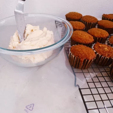 Keto sugar-free cream cheese frosting in a bowl with carrot cupcakes on a plate.