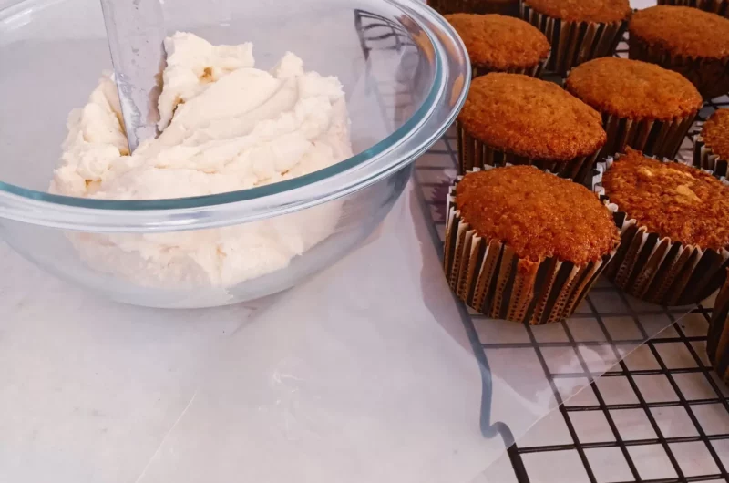 Keto sugar-free cream cheese frosting in a bowl with carrot cupcakes on a plate.