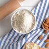 Almond flour ina bowl on a towel wiht a rolling pin and bowl of whole almonds.