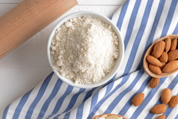 Almond flour ina bowl on a towel wiht a rolling pin and bowl of whole almonds.