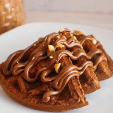 Gluten-free chocolate hazelnut waffles on a plate garnished with sugar-free chocolate hazelnut spread and hazelnuts.