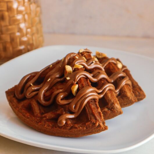 Gluten-free chocolate hazelnut waffles on a plate garnished with sugar-free chocolate hazelnut spread and hazelnuts.
