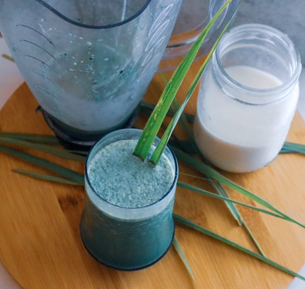 Fever grass green oat milk smoothie in a glass with a blender and oat milk in the background.