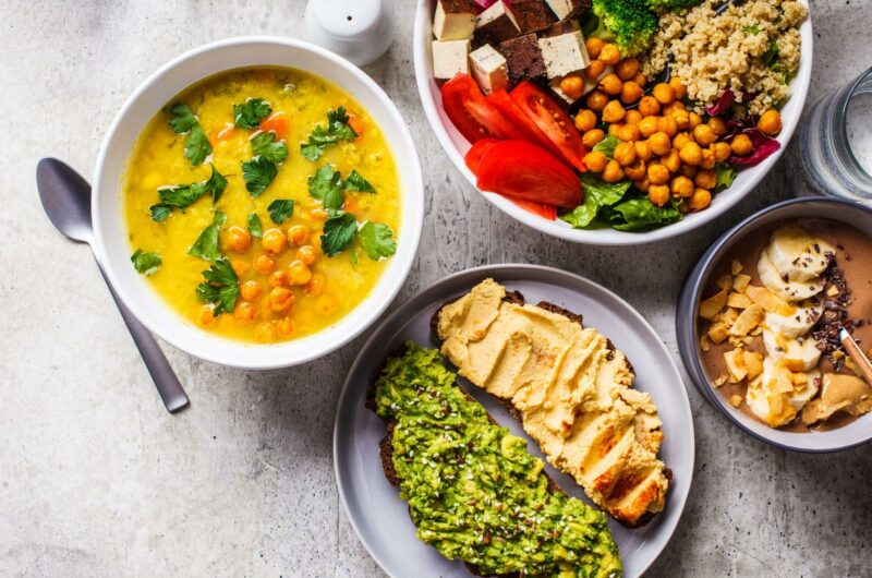 Chocolate smoothie bowl, lentil chickpea soup, tofu Buddha bowl, and toasts arranged on the countertop,