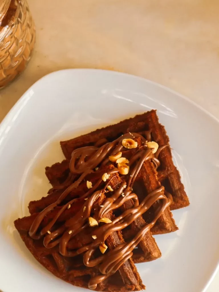 Gluten-free chocolate hazelnut waffles on a plate garnished with sugar-free chocolate hazelnut spread and hazelnuts.